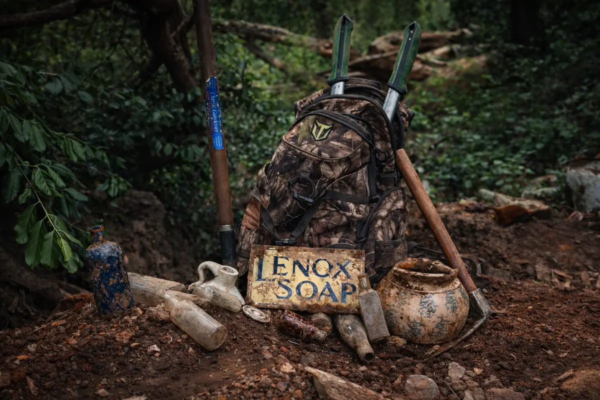 A camouflage backpack, digging tools, a vintage Lenox Soap sign, bottles, and pottery arranged on a forest floor, photographed for KyCreekers.